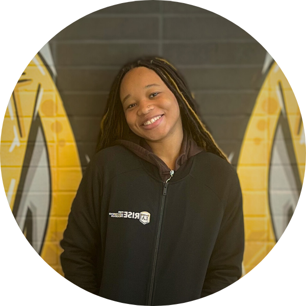 A woman with a black jacket smiling in front of a black wall with yellow markings.