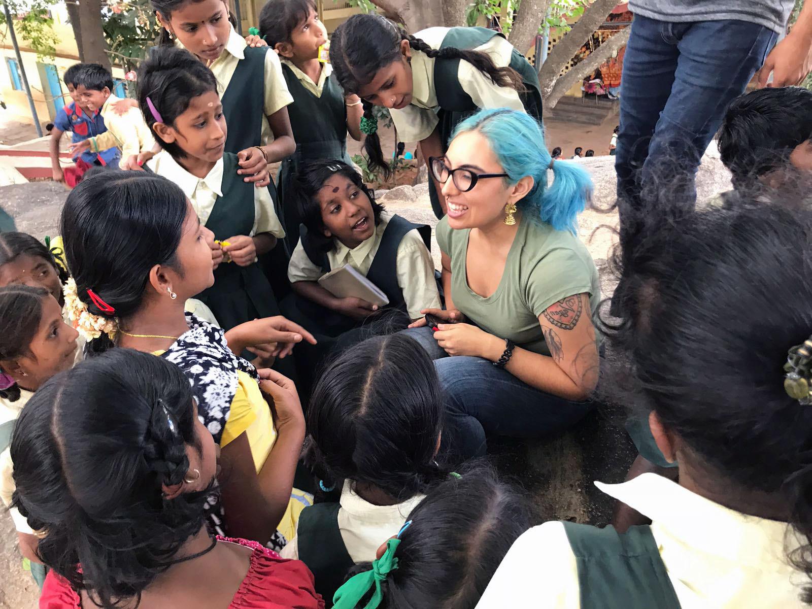 Smiling 加州大学洛杉矶分校 student sitting on the ground surrounded by listening young school students in India.