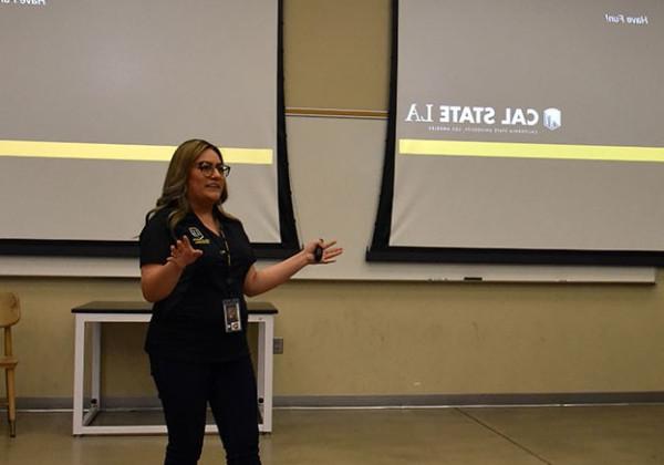 A person standing at the font of a lecture hall in front of two large screens.