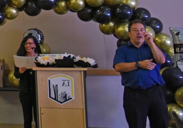 Sign language interpreter standing in the foreground, a person speaking at a podium in the background.