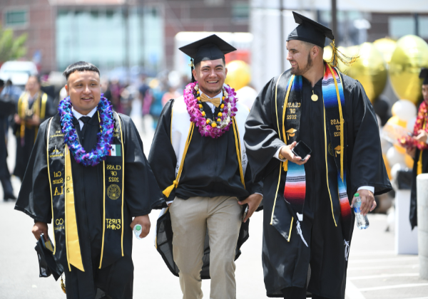 Three male students walking on campus wearing graduation regalia.