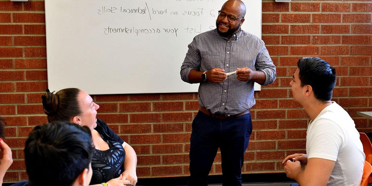 Career advisor stands next to a dry erase board, chatting with students who are seated.