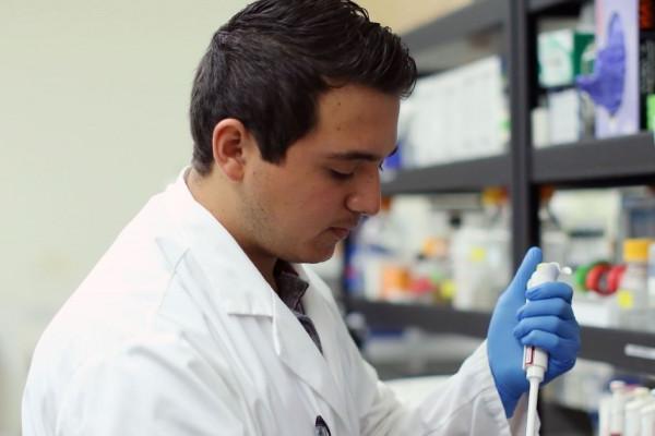 Image of the side profile of a student in a while lab coat and blue gloves using a lab instrument. 