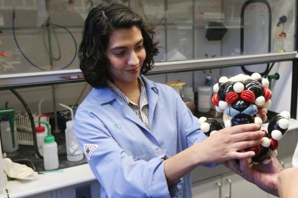 Image of a student in a blue lab coat presenting a model of an atom to a group of fellow students. 