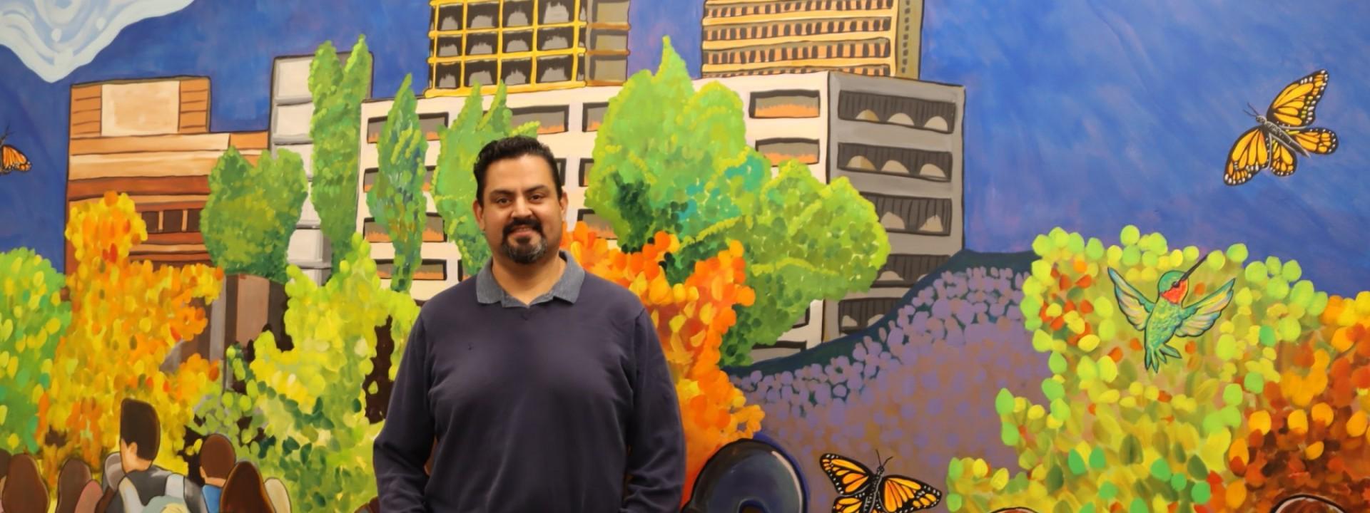 MA Student, Reynaldo Mora posing in front of a mural he painted 