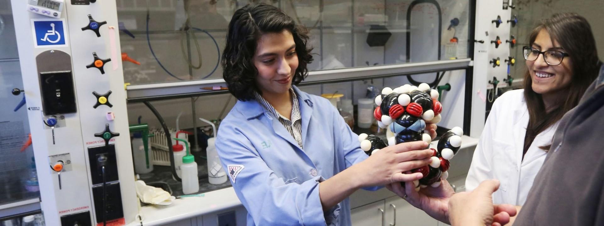 Image of a student in a blue lab coat presenting a model of an atom to a group of fellow students. 