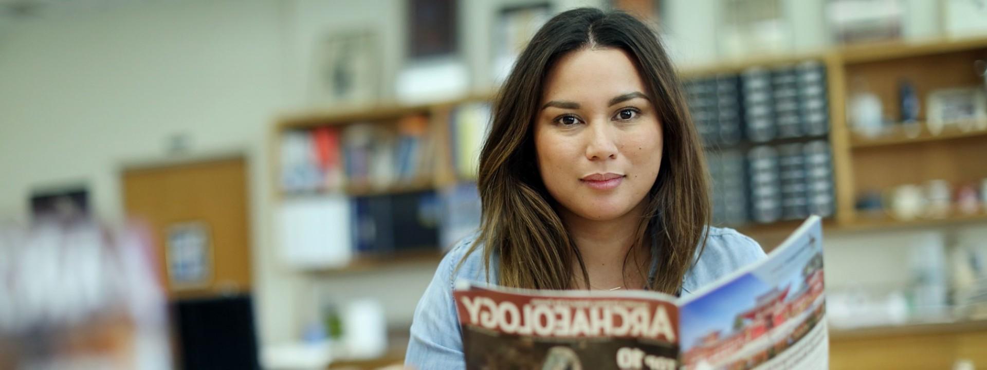 Image of a female student looking directly into the camera as she holds a copy of Archaeology Magazine in a classroom. 